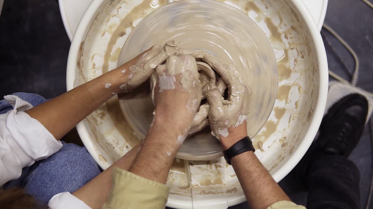Footage from above of dirty hands of romantic couple in love working together on potter wheel and sculpting clay pot, unrecognisable woman and man mold a vase in craft studio workshop. Slow motion