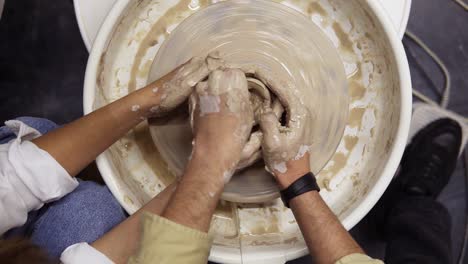 Footage-from-above-of-dirty-hands-of-romantic-couple-in-love-working-together-on-potter-wheel-and-sculpting-clay-pot,-unrecognisable-woman-and-man-mold-a-vase-in-craft-studio-workshop.-Slow-motion