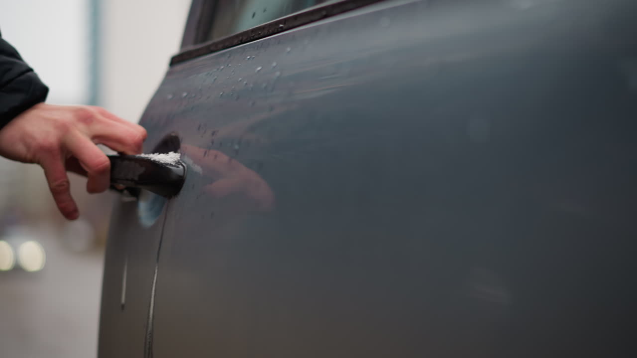 Close up owner opening door handle on ash coloured car with metallic finish, water droplets and snow on surface during winter, hand gripping handle gently beside parked cars on urban street