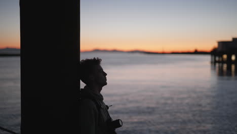 silhouette of young man relaxing with camera on pier, sunset over water