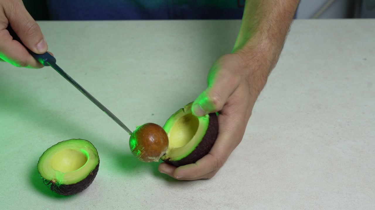 Et nærbillede fra høj vinkel af mandehænder, der bruger en kniv til at fjerne stenen fra en skåret avocado. Madlavningen finder sted på en lys køkkenbordplade, oplyst af levende grønt studiebelysning.