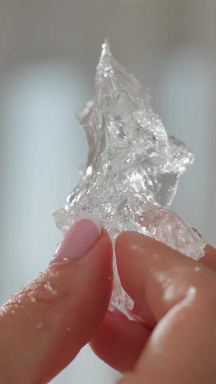 Close up of hand with pink nail polish holding and manipulating clear crystalline sugar glass. subject demonstrates texture detail as they stretch or tear it apart revealing its fragile