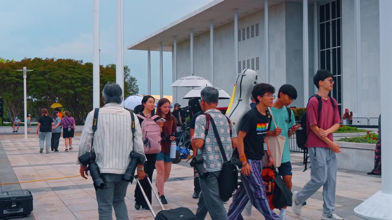 Washington, AS, 13 Juni 2026: Pejalan kaki dan musisi berjalan di dekat pintu masuk Kennedy Center, Washington D.C. Kotak instrumen dan kamera di antara kerumunan di plaza batu yang diterangi matahari dengan kolom-kolom.
