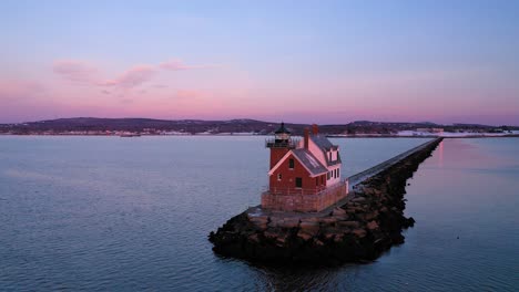 low aerial orbit around a red brick lighthouse at the end of a snow dusted rocky breakwater away from a setting full moon