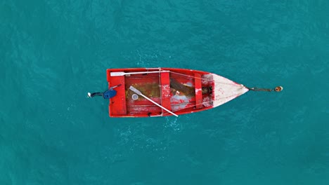 Old-Red-wooden-boat-with-dirty-water-tethered-to-buoy-with-paint-chipping-and-oars-left-inside,-aerial-top-down-overview