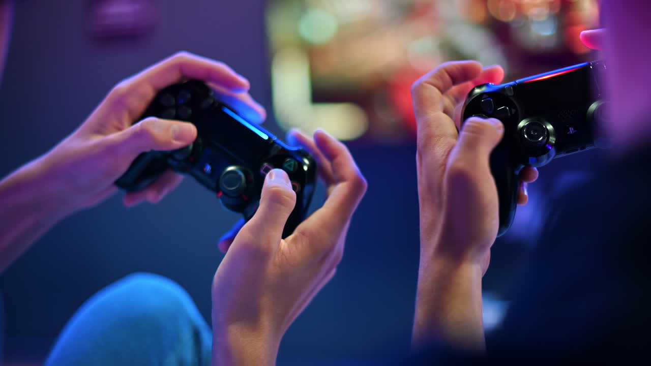 CHISINAU, MOLDOVA - SEPTEMBER 9, 2023: Two teen friends are playing a game console using gamepads, close view of hands, TV on the background. Blue and red illumination