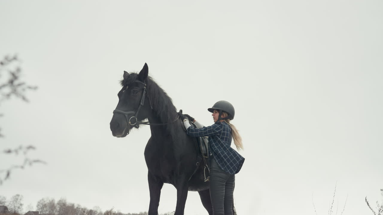 Donna in sella a un alto cavallo nero in un campo nuvoloso. Ritratto composto e dolce abbraccio dopo la cavalcata. La cavallerizza mostra una postura sicura, poi si china per abbracciare il cavallo vicino a cespugli spogli. Orizzonte rurale tranquillo e colori tenui.