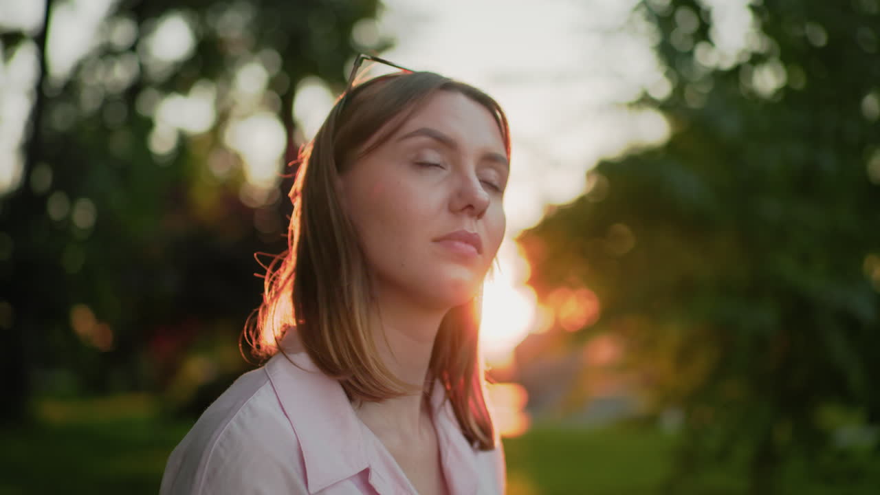 joven con camiseta rosa con gafas en la cabeza, con una cálida sonrisa, mira hacia abajo pensativa, bañada en la luz del sol, con llamaradas solares y vegetación borrosa en el fondo