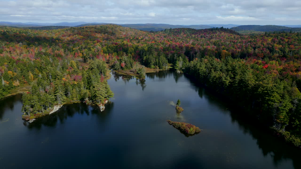 Aerial pullback over dark lake reflecting clouds in sky on calm water as trees begin to change colors and cloud shadows pass over top