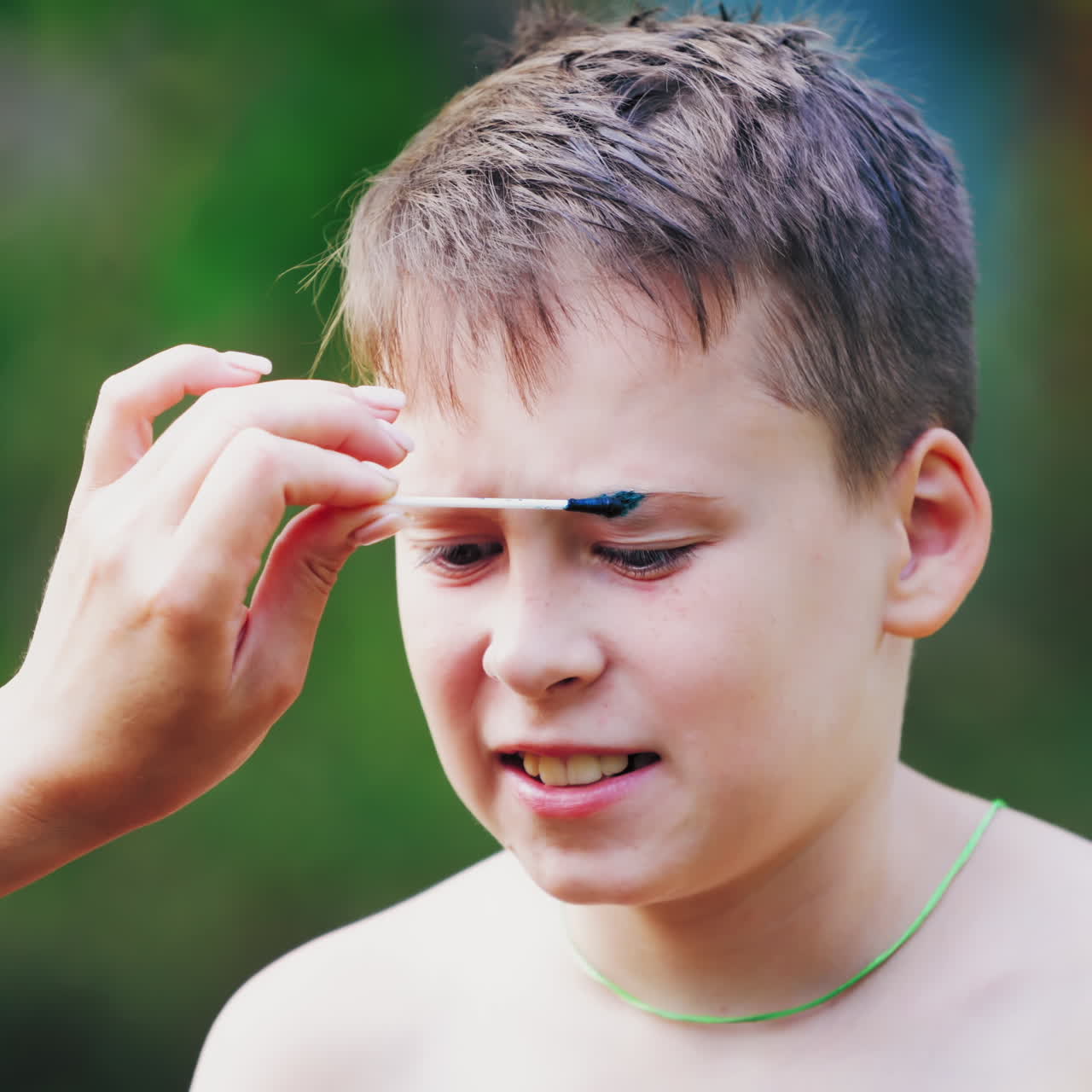 Boy has a open wound on the eyebrow. Mother gives the first aid to her child on face. Naked boy has an injury on his eyebrow and woman's hand treats it outdoors.