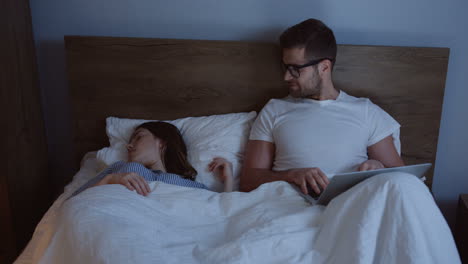 handsome young man in pajama and glasses working late at night on the laptop computer while his wife sleeping peacefully near him in the bed