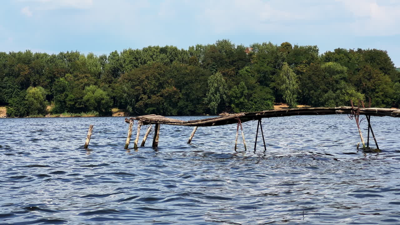 Old unsecure wooden bridge above the river. Waterfront with green trees growing at backdrop.