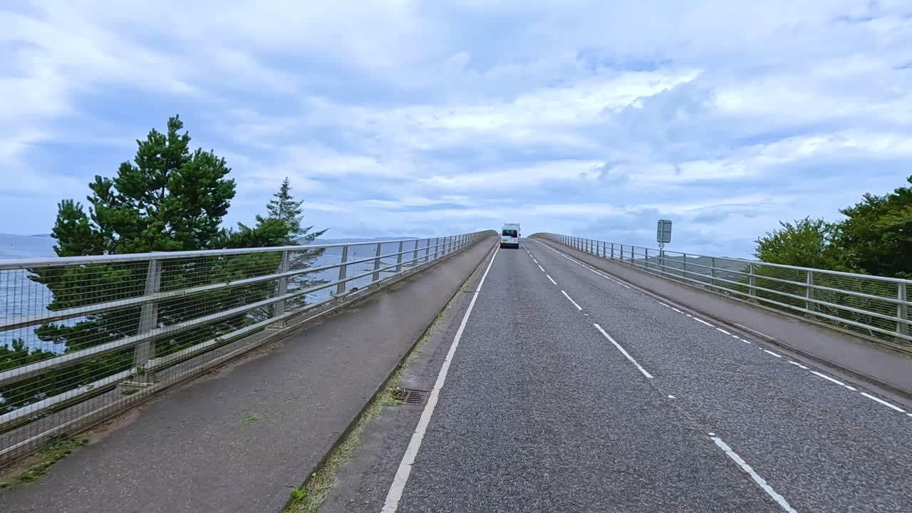 A vehicle travels along a two-lane bridge road with metal railings, surrounded by lush greenery and expansive water views under bright daylight and partly cloudy skies