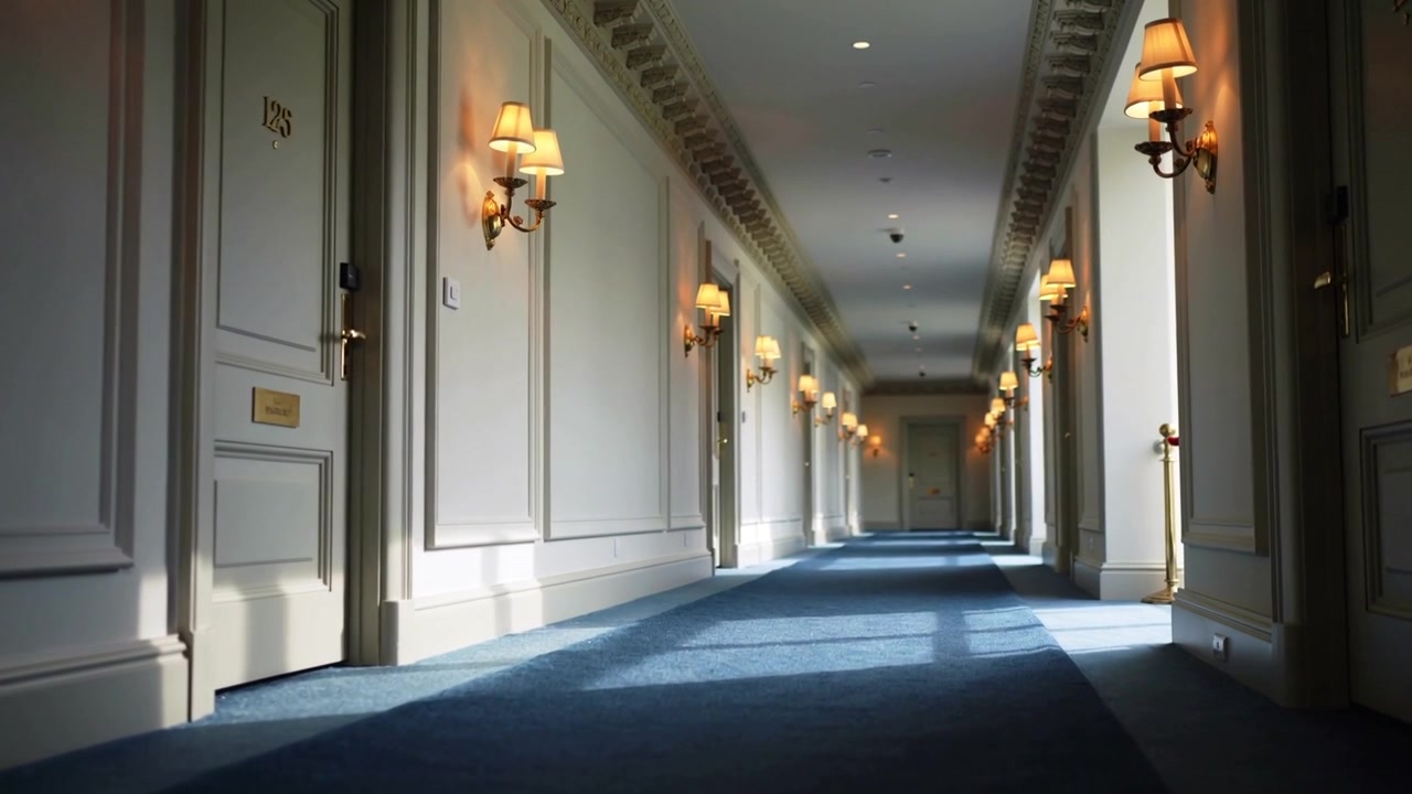 Telecamera a bassa angolazione che si sposta lungo il corridoio di un hotel, rivelando porte con numeri in ottone e applique.