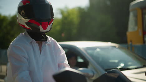 a female motorcyclist, wearing a helmet, is waiting for traffic, leaning slightly backward, in the background, a blurred car is seen with people inside, slowing down due to traffic