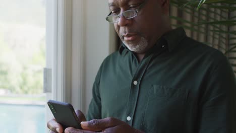 Hombre-Mayor-Afroamericano-Usando-Un-Teléfono-Inteligente-Cerca-De-La-Ventana-En-Casa