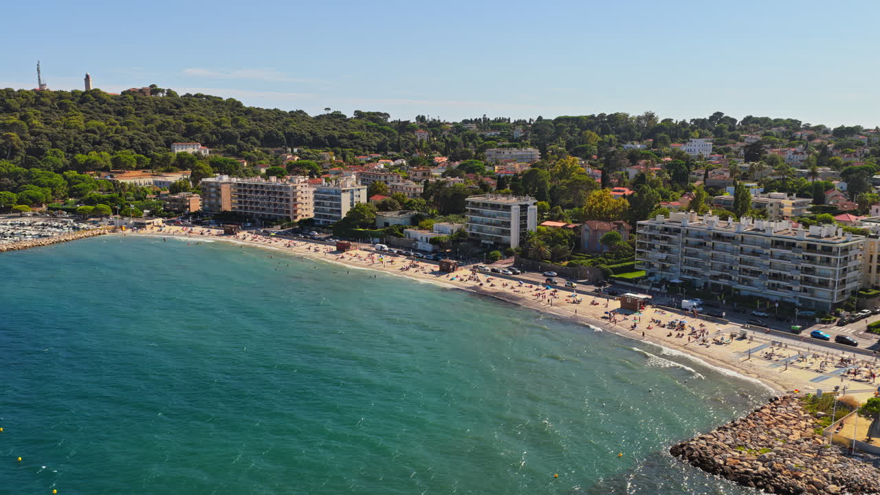 Aerial drone view of Antibes sandy beach filled with sunbathers and swimmers, with seafront apartment buildings and green hills in the background