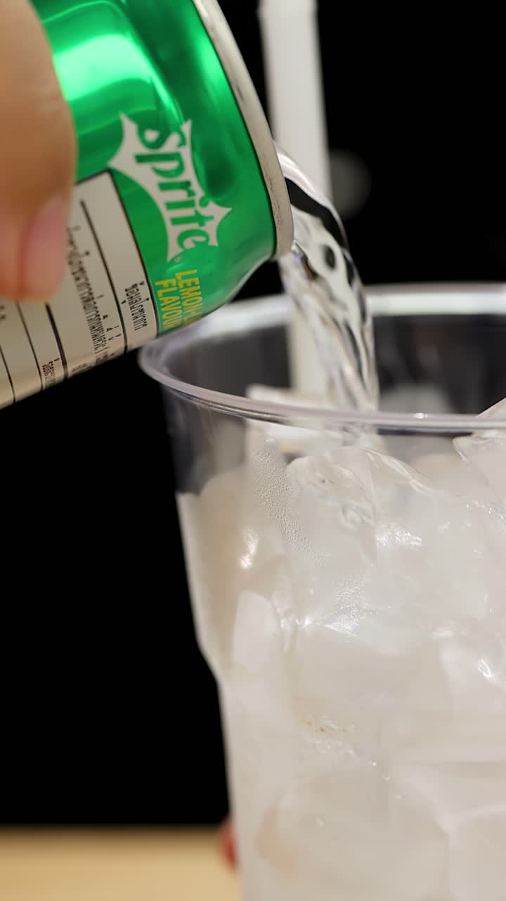 Mão derrama refrigerante claro e efervescente de uma lata em um copo plástico cheio de cubos de gelo.