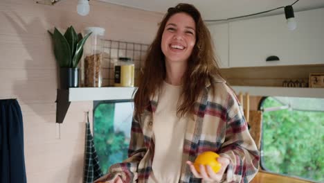 happy brunette girl in a plaid shirt juggles two oranges and dances in her motorhome or trailer outside the city in the summer at a camp on a picnic. happy girl juggling fruits and having fun in her house in summer