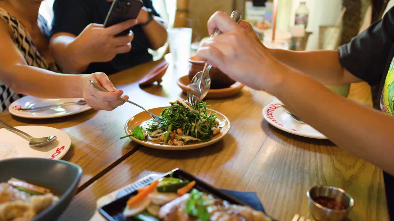Diners serve spicy water mimosa salad at a well-lit Bangkok restaurant, handheld camera, natural lighting