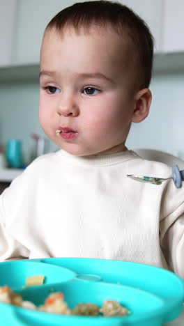 dinner time for a cute little kid. baby sits at feeding table eating from spoon. close up. blurred backdrop. vertical video
