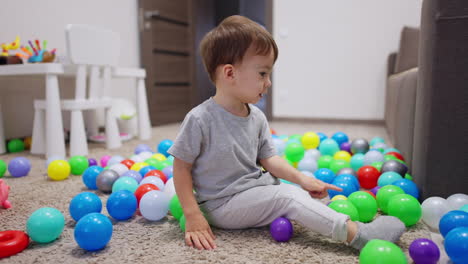 two year old toddler sits on the floor among the colorful balls. kid takes green ball and throws it ahead.