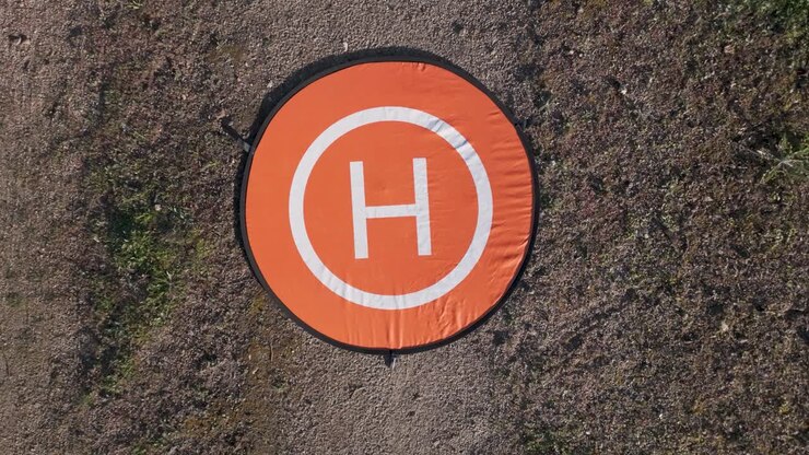 Riprese con un drone con inquadratura zenitale in volo ascendente dove si vede la sua piattaforma di decollo mobile arancione con un bordo nero, in bianco c'è un cerchio interno e una H, il terreno circostante.