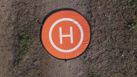 filmando con un dron con una vista de arriba hacia abajo en vuelo ascendente donde vemos su plataforma móvil de despegue en naranja con un borde negro, en blanco hay un círculo interior y una h, el exterior es tierra
