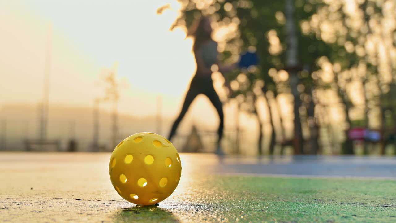 Yellow ball on a blue and green court with a woman playing pickleball at sunrise, after rain on the background