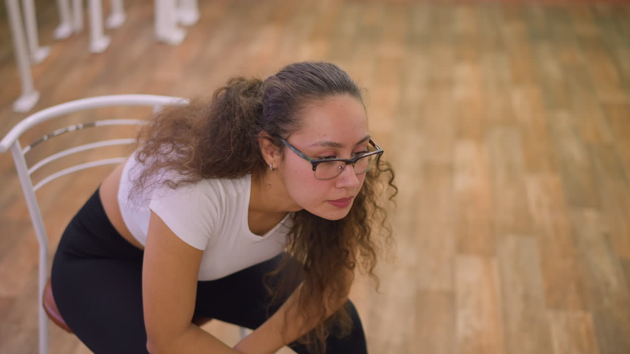 Close view of woman in white top and black leggings sitting on chair breathing heavy with head down, looking tired and exhausted after workout in bright studio with wooden floor and soft light