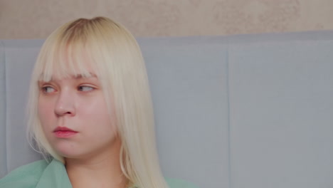 close up of blonde girl chewing food with neutral facial expression while sitting indoors, pale blue headboard and beige wall create calm setting