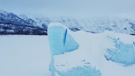 Large-blue-ice-boulder-stands-tall-in-middle-of-snowy-Alaskan-landscape,-aerial