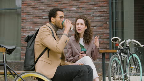 young american man and woman in formal clothes drinking coffee and talking while sitting next to their bikes on a wooden bench in the city