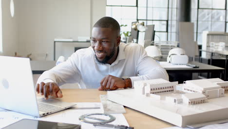 smiling african american businessman working on laptop with architectural model in office