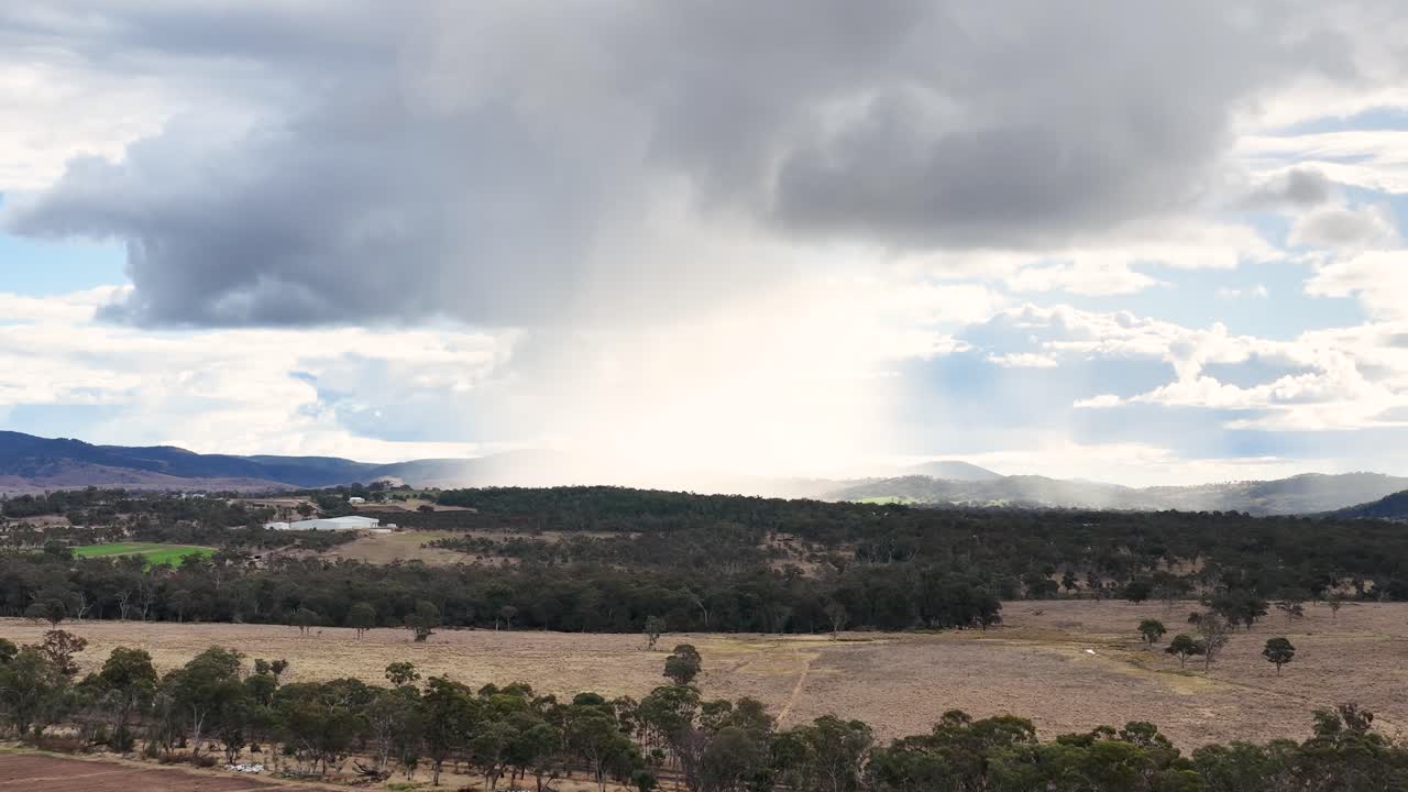 A distant rain shower drifts over open farmland and forested hills under dramatic clouds, captured in a wide, steady shot with natural daylight