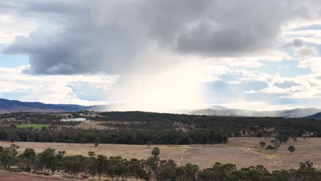 a distant rain shower drifts over open farmland and forested hills under dramatic clouds, captured in a wide, steady shot with natural daylight