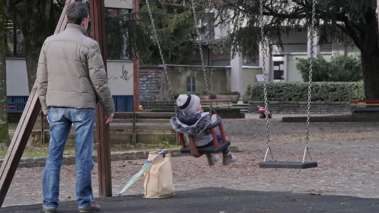 A Masked Man Pushes His Little Daughter on the Swing at the Park in Town. During Winter. Milan, Italy