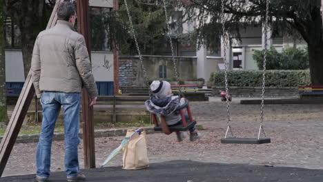 a masked man pushes his little daughter on the swing at the park in town. during winter. milan, italy