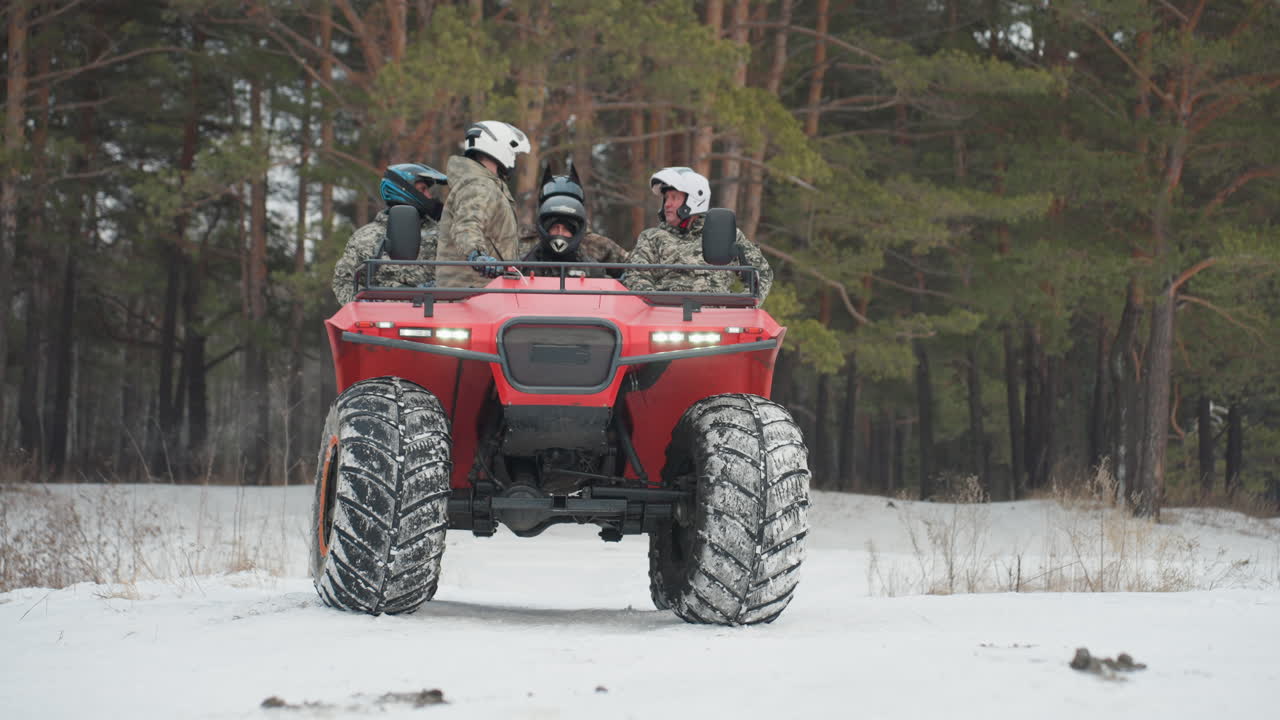 Vista frontale di un quad rosso sulla neve, con due piloti caschi e abbigliamento mimetico seduti pronti sul robusto quad con pneumatici chiodati. Sullo sfondo una pineta e luce invernale nuvolosa, che crea una tesa attesa prima dell'accelerazione.