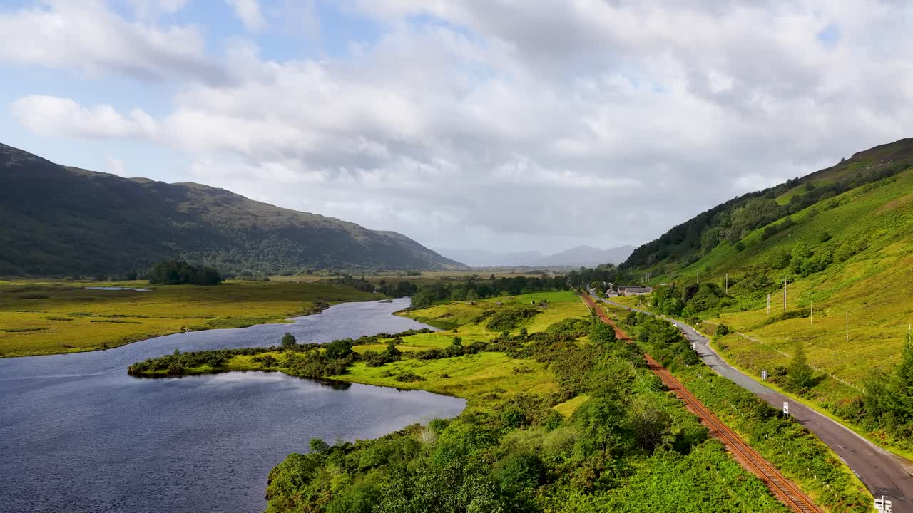 Drone glides above serene lake, green valley, and winding road under soft daylight in Scotland