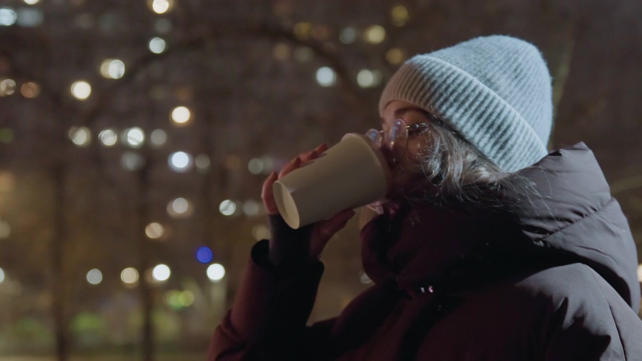 Donna al parco di notte che sorseggia caffè, avvolta in un piumino, con berretto e occhiali sotto il bokeh soffuso del lampione, sguardo contemplativo verso lo skyline, respiro lento visibile nell'aria fredda, mani che scaldano la tazza