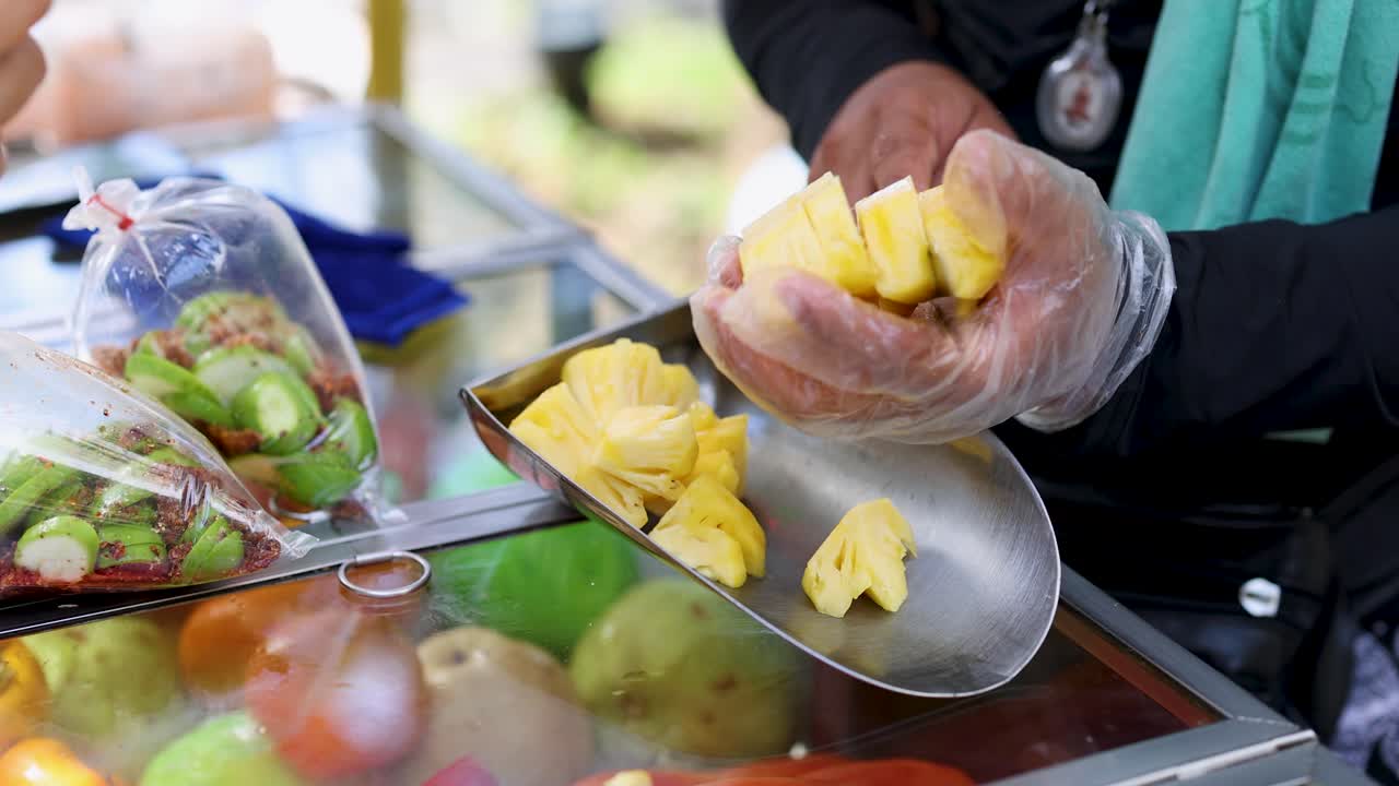 Gloved vendor slices fresh pineapple at outdoor fruit cart, natural daylight, close-up handheld shot