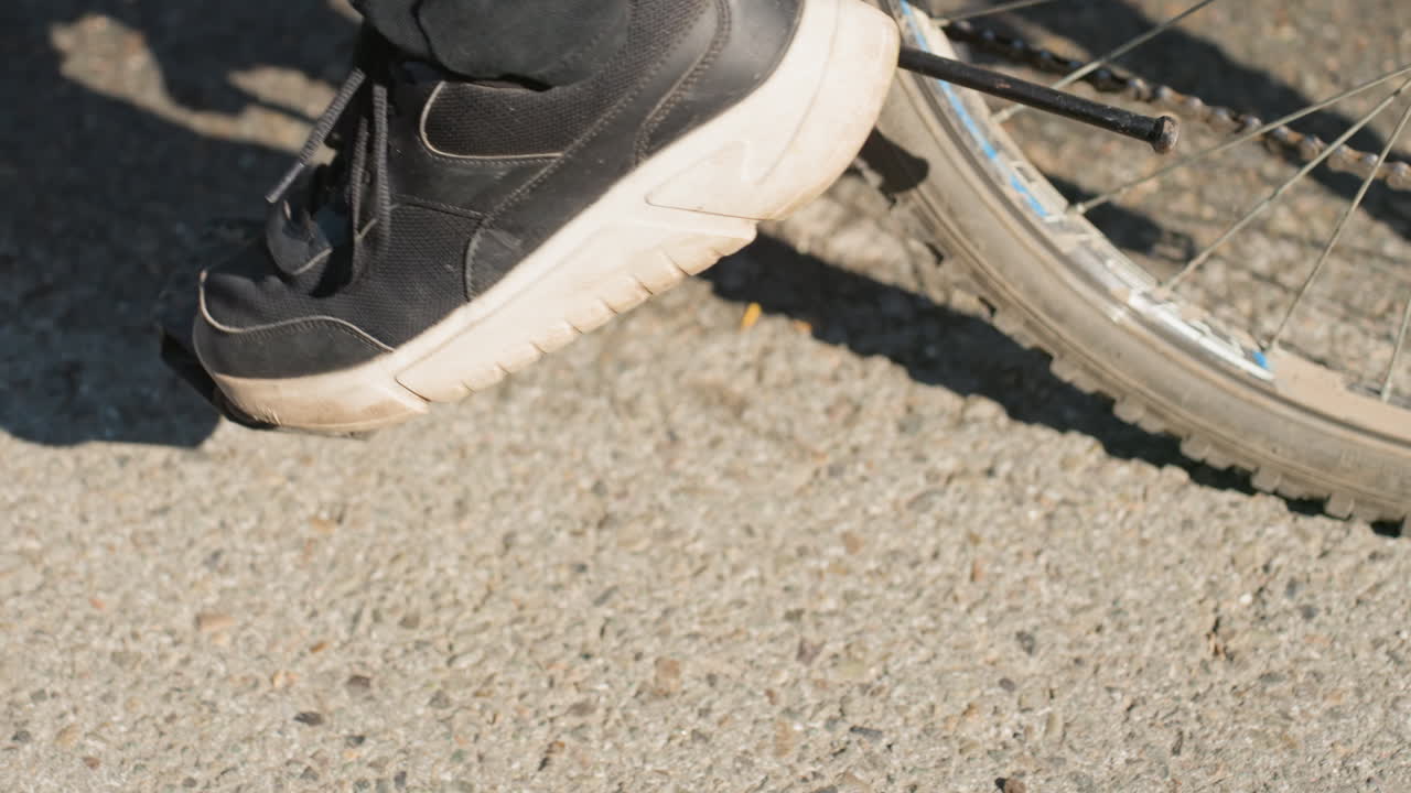 ペダルに足をかけたバイク便の足元のクローズアップ。使い古されたスニーカーが埃っぽいペダルに押し付けられている。配達の合間に休憩する都市部の配達員。フレーム外には小包バッグが見える。太陽光が長く影を落とし、アスファルトの質感が見える。
