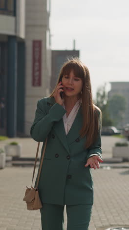 annoyed young woman in business pantsuit shouts into mobile phone walking down street on rainy day. irritated female person speaks waving hand angrily