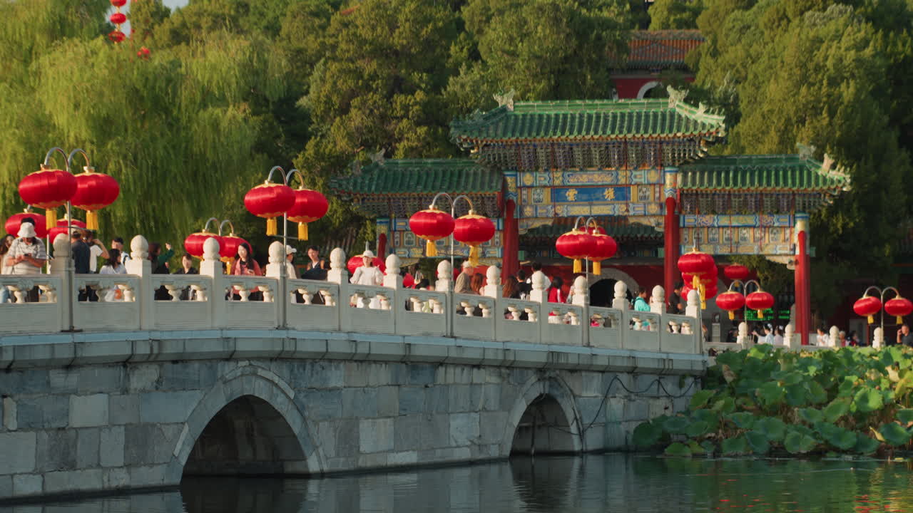 Ponte di pietra fiancheggiato da lanterne rosse e una folla vivace, con una balaustra ornata sopra uno stagno di fiori di loto. Bagliore serale e riflessi, famiglie e turisti che passeggiano, venditori ambulanti e un fotografo che cattura la scena.