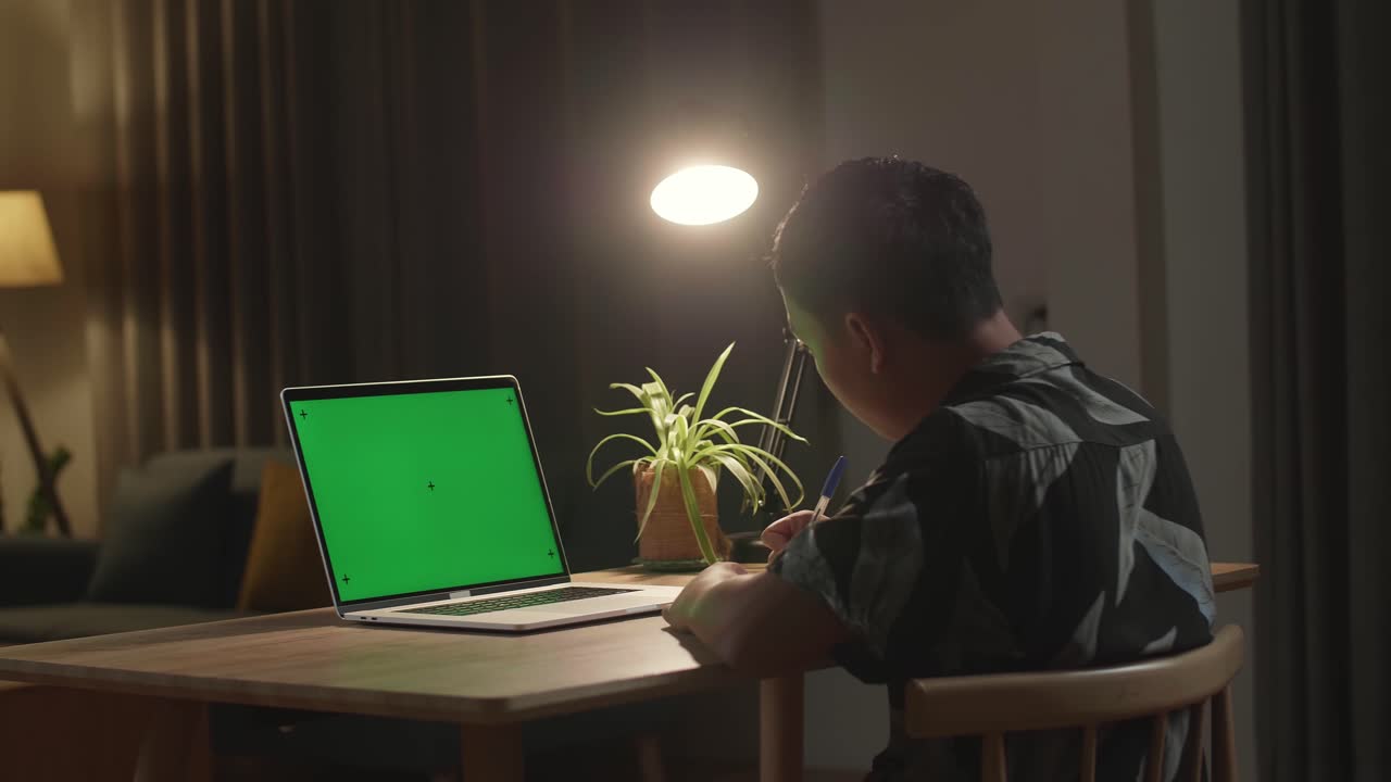 Teenage Boy Sitting At His Desk Learning Online On A Laptop With Green Mock-Up Screen. Late At Night In His Living Room Boy Uses Notebook Computer And Writing On Book