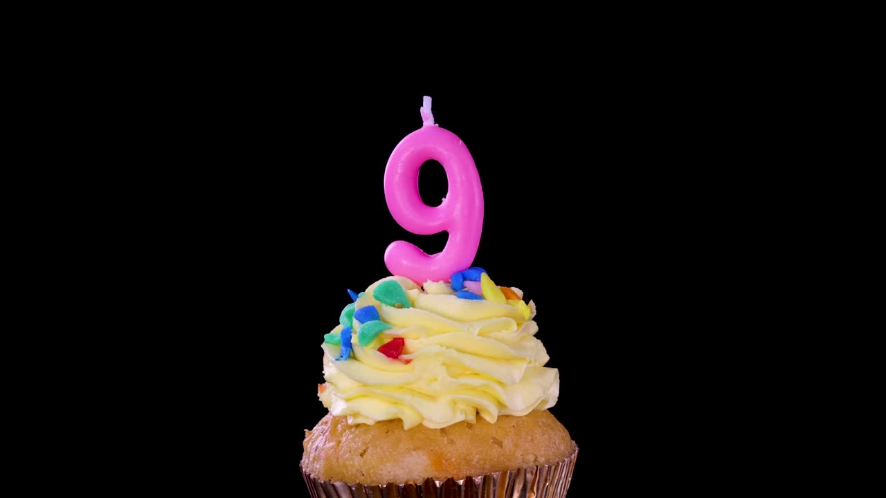 Hand inserts pink number nine birthday candle into yellow frosted cupcake with sprinkles, black background