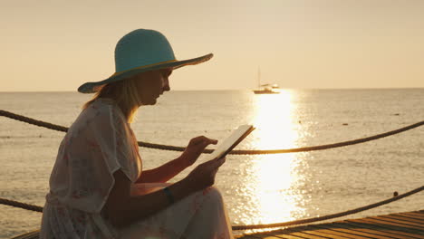 Un-Turista-Con-Una-Tableta-Se-Sienta-En-El-Muelle-Pintoresco-Amanecer-Sobre-El-Mar-En-El-Fondo-Video-4k