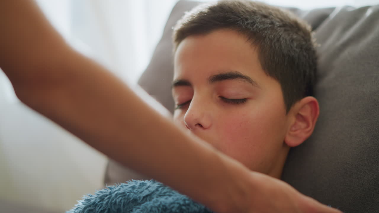 Close-up view of a caring hand covering a sleeping boy with a soft blue blanket, light filters through the window, creating a peaceful and intimate moment