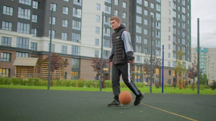 Young white man dribbling basketball outdoors, walking toward hoop on residential court, wearing hoodie and trackpants, carrying ball under overcast sky, apartment towers and fenced playground